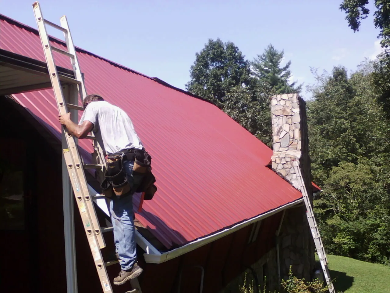 Roofer on a ladder beside a red metal roof and stone chimney — Soffit & Fascia Repair by Roofing Brothers in Port Orchard